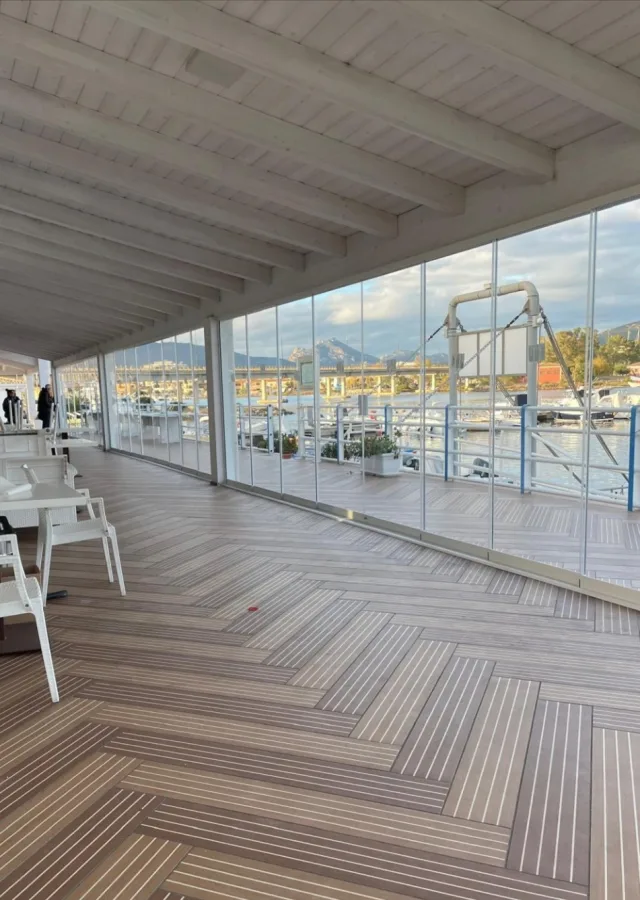 Interno di un dehors vetrato con soffitto a travi di legno chiaro, affacciato su un porto turistico
