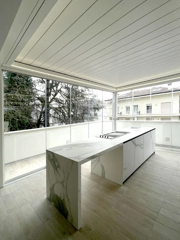 Cucina esterna con isola in materiale effetto marmo, sotto la copertura a lame e accanto alle vetrate