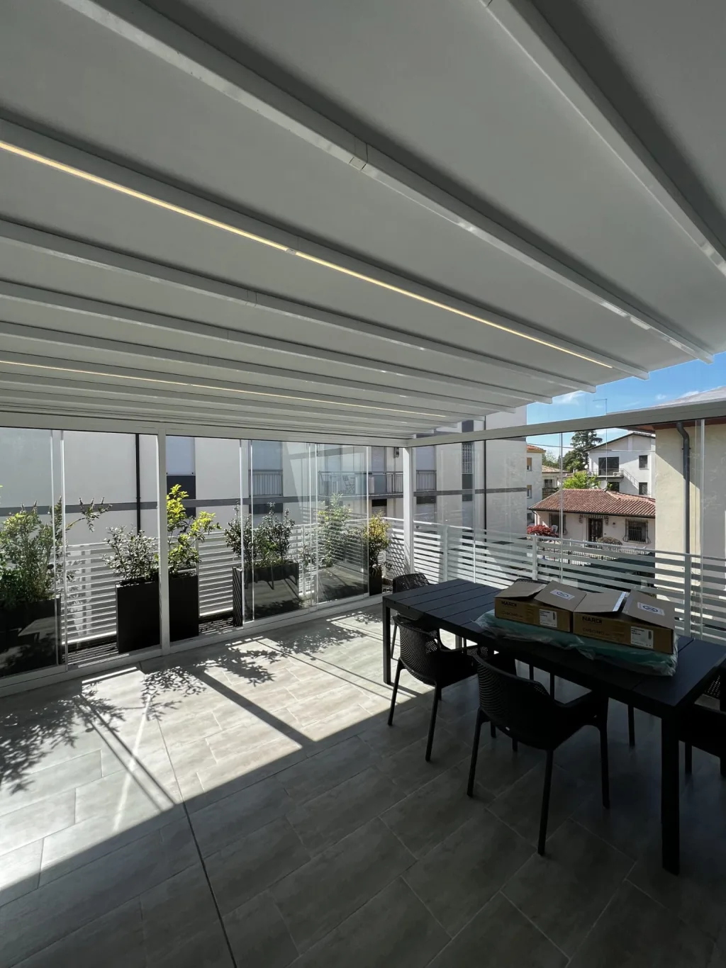 Terrazzo coperto da una pergola bioclimatica bianca con lame orientabili e strisce LED, chiuso su due lati da vetrate scorrevoli trasparenti