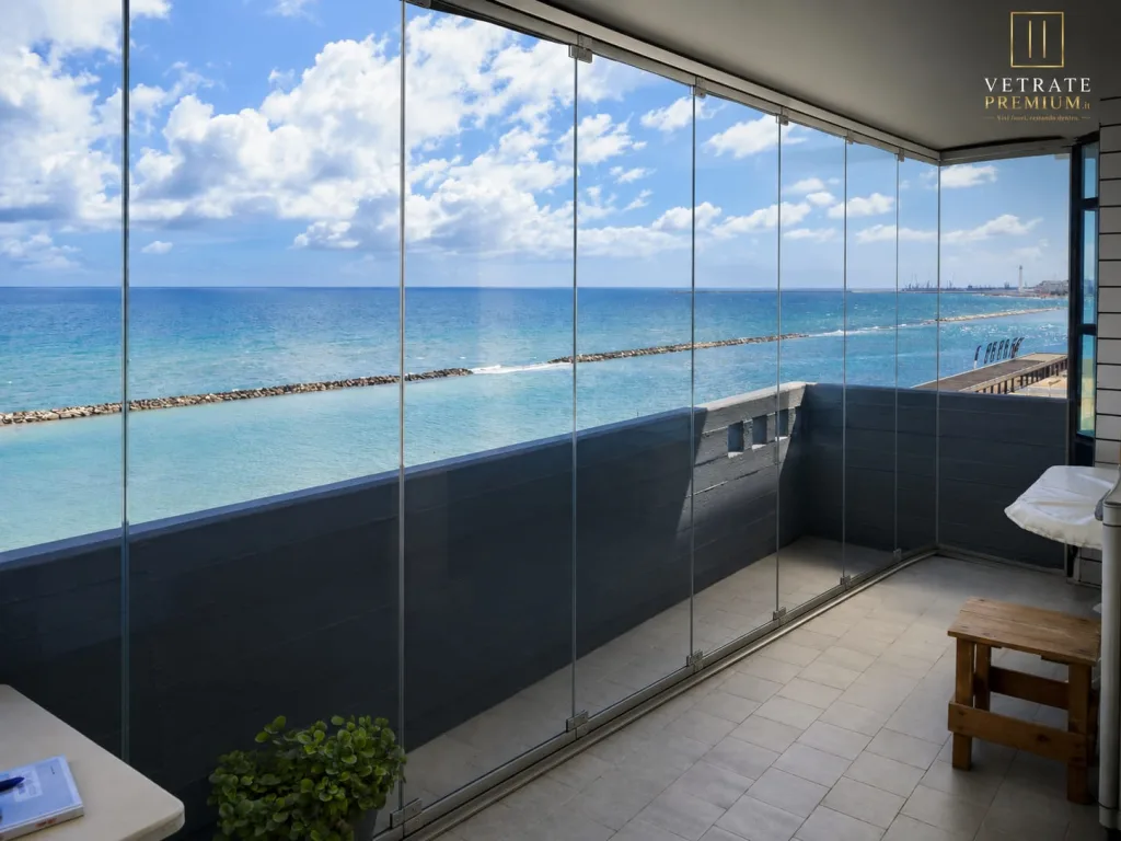 Balcone fronte mare chiuso con vetrate a tutta altezza, con frangiflutti e acqua turchese oltre il vetro